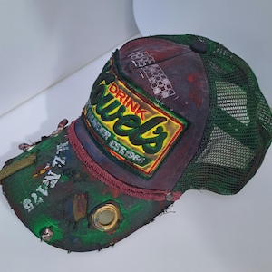Könnte beinhalten: Distressed Trucker-Cap mit grünem Mesh-Rücken und Patchwork-Design. Auf der Vorderseite befindet sich ein Aufnäher mit den Worten "Drink Jewels" und "Thunder Est. 1964". Der Schirm ist grün mit dem Text "L.N. 175".