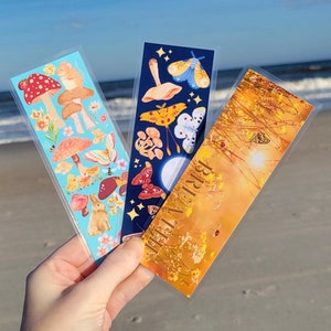 May include: Three bookmarks with different designs. The first bookmark has a blue background with mushrooms, butterflies, and a rabbit. The second bookmark has a dark blue background with mushrooms, butterflies, and a moon. The third bookmark has a yellow background with a sunset, flowers, and butterflies. The text "BRIAR THORN" is printed on the third bookmark.