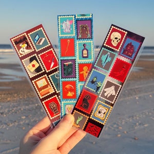 May include: Three colorful bookmarks with a variety of spooky and gothic themed illustrations. Each bookmark is made up of a grid of small squares, each containing a different image. The images include a skull, a ghost, a haunted house, a witch, a spider, a coffin, a potion bottle, a book, a mushroom, a voodoo doll, a crow, a hand reaching out from the ground, and more.