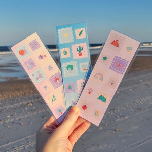 May include: Three colorful bookmark sets with various cute illustrations, including a snail, a fish, a mushroom, a cactus, a tree, a rainbow, a mountain, a butterfly, a bee, and a ladybug. The bookmarks are on a white sandy beach.