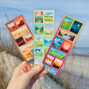 May include: Three sticker sheets with landscape illustrations are held up against a beach and sky backdrop. Each sheet features multiple rectangular stickers with images of mountains, deserts, and bodies of water. The sheets are in clear plastic.