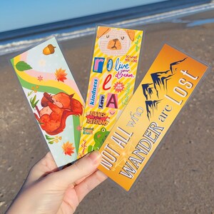 May include: Three colorful bookmarks with inspirational quotes. The first bookmark features a squirrel holding an acorn, the second bookmark has a dog with the words "Smile, Live Your Dream", and the third bookmark has a mountain range with the words "Not All Who Wander Are Lost".