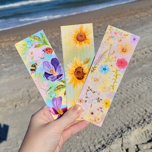 May include: Three floral bookmarks are held up against a beach backdrop. Each bookmark features a different floral design, including sunflowers, purple crocus, and a variety of other flowers. The bookmarks are colorful and detailed, with a watercolor style.