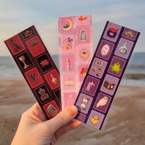 May include: Three rectangular bookmarks featuring grid-style illustrations. The left bookmark has a dark red background with icons of a cloak, chalice, rose, and throne. The center is light pink with a moon, tower, and wand. The right is purple with a cauldron, potion, frog, and owl.