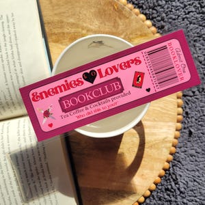 May include: Pink and black bookmark with the text "Enemies to Lovers Bookclub" and "Tea Coffee & Cocktails provided" with a small illustration of a rose and a dagger. The bookmark also has a barcode and the text "Admit One Booklover".