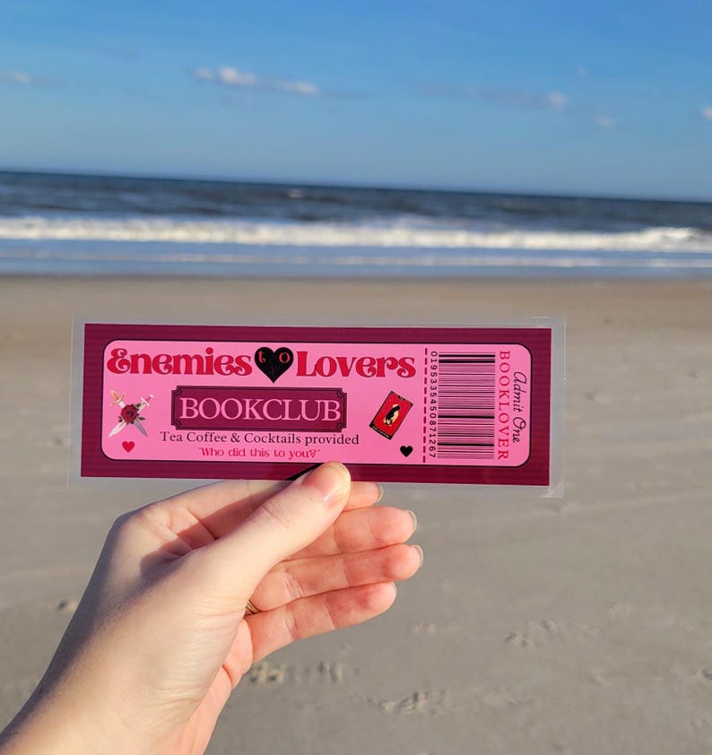 May include: A pink and red bookmark with the text "Enemies Lovers Bookclub" and the text "Tea Coffee & Cocktails provided" and "Who did this to you?"  The bookmark has a barcode and the text "Admit One BOOKLOVER".