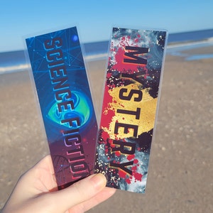 May include: Two bookmarks with colorful designs. One bookmark has a blue and purple galaxy design with the text "Science Fiction" and the other bookmark has a red and black design with the text "Mystery".