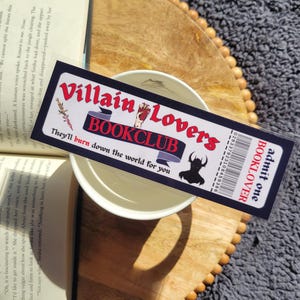 May include: A black and red bookmark with the text "Villain Lovers Bookclub" and "They'll burn down the world for you". The bookmark also has a barcode and the text "admit one BOOKLOVER".