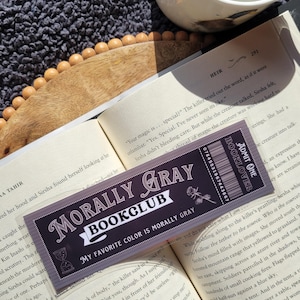 May include: A close-up of a book club bookmark with the text "Morally Gray Bookclub" and the tagline "My favorite color is morally gray". The bookmark is made of clear plastic with a black and white design. The bookmark is placed on top of an open book.