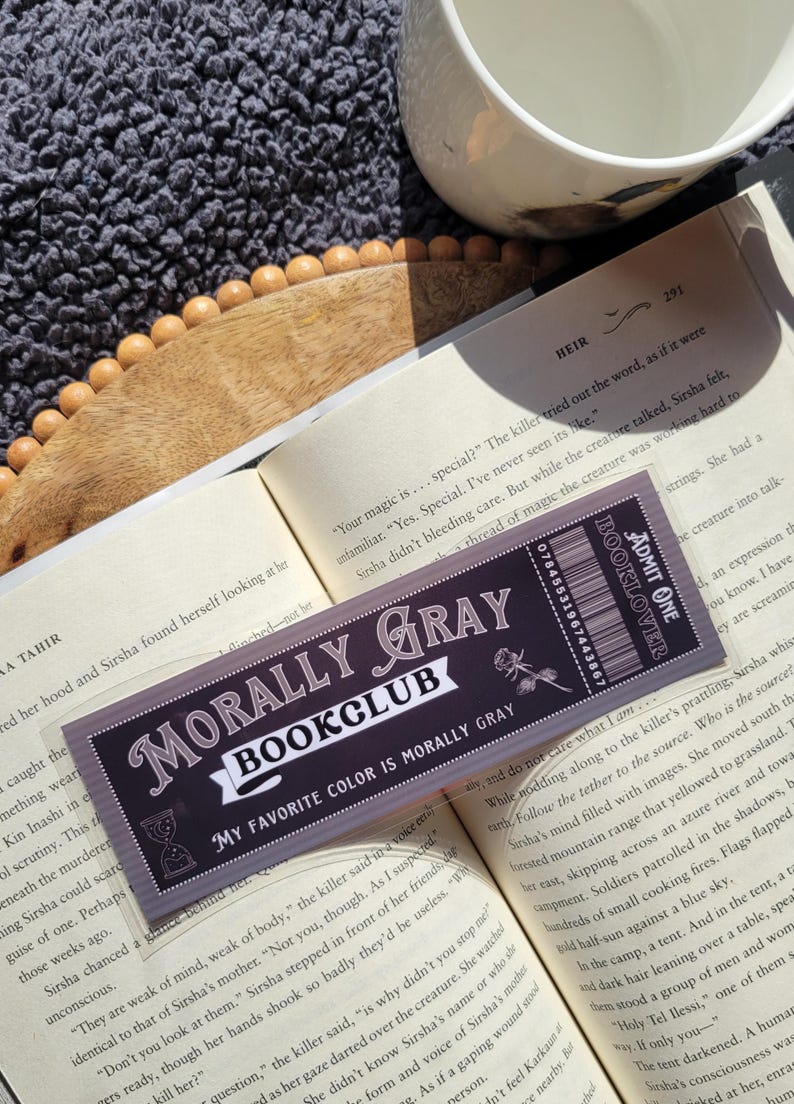 May include: A close-up of a book club bookmark with the text "Morally Gray Bookclub" and the tagline "My favorite color is morally gray". The bookmark is made of clear plastic with a black and white design. The bookmark is placed on top of an open book.