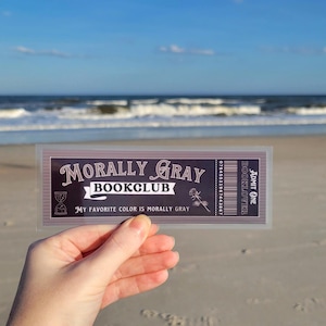 May include: A ticket for the "Morally Gray Bookclub" with the text "My favorite color is morally gray" and a rose illustration. The ticket is held in a hand against a beach background.
