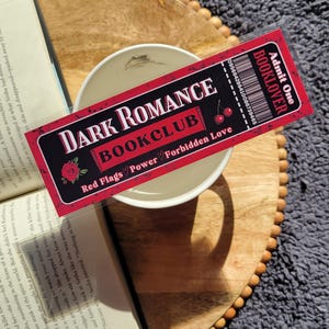 May include: A red and black bookmark with the text "Dark Romance Bookclub" and "Red Flags Power Forbidden Love". The bookmark is shaped like a ticket with a cherry graphic and the text "Booklover" and "Admit One".