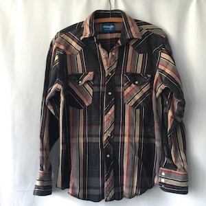 May include: A long-sleeved Wrangler shirt with a plaid pattern in black, tan, red, and white stripes. The shirt has two chest pockets with button closures and a button-down front. The collar is a classic spread style.