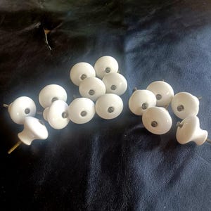 May include: A collection of white ceramic cabinet knobs with brass screws. The knobs are round with a glossy finish, and are scattered on a dark surface. These knobs are suitable for furniture restoration or new builds.