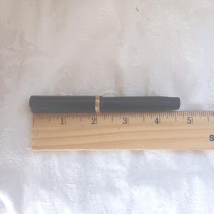 K&ouml;nnte beinhalten: Ein schwarzer F&uuml;llfederhalter mit goldenem Band, auf einem Holzlineal. Der Stift ist etwa 13 cm lang. Das Lineal ist in Zoll unterteilt.