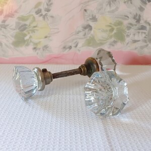 May include: Two clear, faceted glass door knobs with a multi-sided, star-like shape. The knobs are connected by a central metal spindle with a threaded surface and a small set screw, featuring a metallic base plate behind each glass knob.