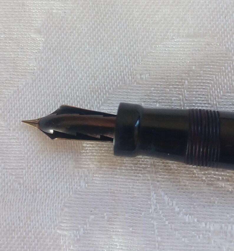 K&ouml;nnte beinhalten: Nahaufnahme eines schwarzen F&uuml;llfederhalters mit goldener Feder. Der Stiftk&ouml;rper ist schwarz mit einem gerippten Design in der N&auml;he der Spitze. Die Feder ist goldfarben und spitz, zum Schreiben bestimmt. Der Stift befindet sich vor einem wei&szlig;en, strukturierten Hintergrund.