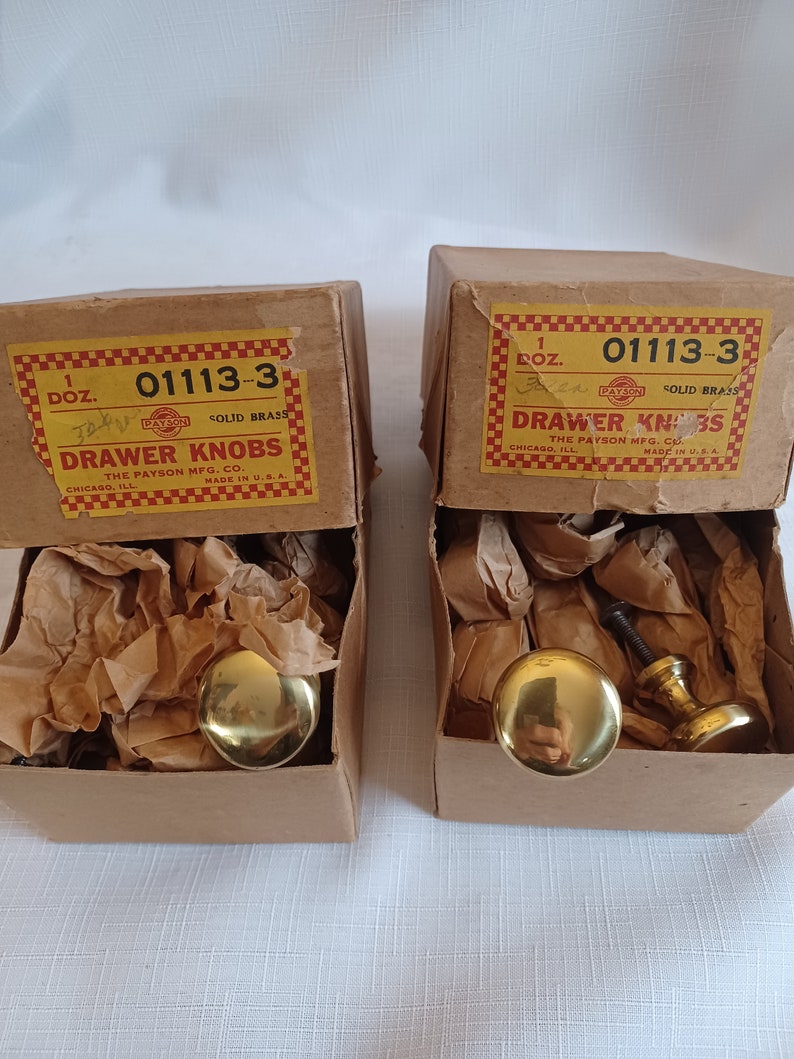 Może przedstawiać: Dwa pudełka z mosiężnymi gałkami do szuflad. Pudełka są oznaczone jako "DRAWER KNOBS" i "THE PAYSON MFG. CO." Gałki są w kolorze złotym i mają zaokrąglony kształt. Wyprodukowano w USA.