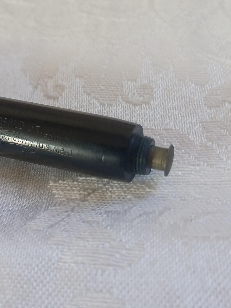 K&ouml;nnte beinhalten: Nahaufnahme eines schwarzen F&uuml;llfederhalters mit goldfarbenen Akzenten. Der Stift hat Text an der Seite und eine aufschraubbare Spitze. Der Stift liegt auf einer wei&szlig;en, strukturierten Oberfl&auml;che.