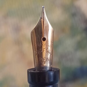 K&ouml;nnte beinhalten: Nahaufnahme einer goldfarbenen F&uuml;llfederhalterfeder mit dem Markennamen "PARKER" und dem Wort "PEN". Die Feder hat eine feine Spitze und ist vor einem verschwommenen Hintergrund platziert. Der Stiftk&ouml;rper ist schwarz.