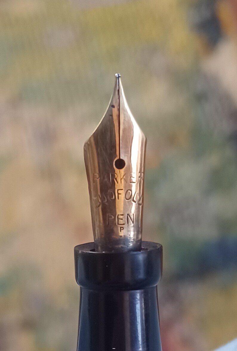 K&ouml;nnte beinhalten: Nahaufnahme einer goldfarbenen F&uuml;llfederhalterfeder mit dem Markennamen "PARKER" und dem Wort "PEN". Die Feder hat eine feine Spitze und ist vor einem verschwommenen Hintergrund platziert. Der Stiftk&ouml;rper ist schwarz.