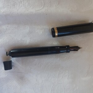 K&ouml;nnte beinhalten: Ein schwarzer F&uuml;llfederhalter mit einem goldenen Band am Deckel und einer dunklen Feder. Der Stift ist zerlegt, mit Kappe und einem kleinen Teil getrennt vom Hauptk&ouml;rper. Der Stift liegt auf einer wei&szlig;en, strukturierten Oberfl&auml;che.