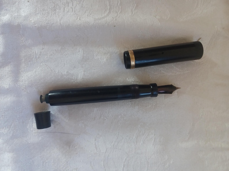 K&ouml;nnte beinhalten: Ein schwarzer F&uuml;llfederhalter mit einem goldenen Band am Deckel und einer dunklen Feder. Der Stift ist zerlegt, mit Kappe und einem kleinen Teil getrennt vom Hauptk&ouml;rper. Der Stift liegt auf einer wei&szlig;en, strukturierten Oberfl&auml;che.