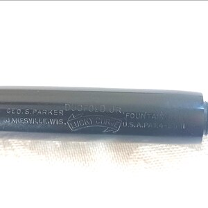 K&ouml;nnte beinhalten: Nahaufnahme eines schwarzen F&uuml;llfederhalters mit silbernem Text. Der Stift ist mit "DUOFOLD JR. FOUNTAIN" und "LUCKY CURVE" sowie dem Namen Geo. S. Parker und dem Standort Janesville, Wis. gekennzeichnet. Der Stift hat eine Schraubkappe.