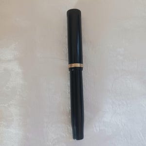 K&ouml;nnte beinhalten: Ein schwarzer Stift mit einem goldenen Band in der Mitte. Der Stift hat eine zylindrische Form und scheint aus einem glatten, gl&auml;nzenden Material zu bestehen. Er liegt auf einer wei&szlig;en, strukturierten Oberfl&auml;che.