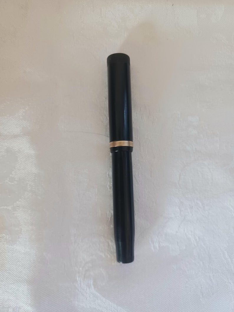 K&ouml;nnte beinhalten: Ein schwarzer Stift mit einem goldenen Band in der Mitte. Der Stift hat eine zylindrische Form und scheint aus einem glatten, gl&auml;nzenden Material zu bestehen. Er liegt auf einer wei&szlig;en, strukturierten Oberfl&auml;che.