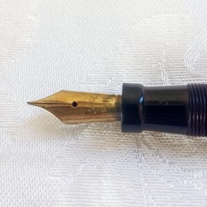 K&ouml;nnte beinhalten: Nahaufnahme einer Feder eines F&uuml;llfederhalters mit goldfarbener Spitze und schwarzem Schaft. Die Feder hat eingravierten Text und ein kleines Loch. Der Stift befindet sich vor einem wei&szlig;en, strukturierten Hintergrund.