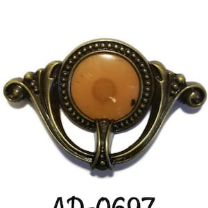 May include: Antique bronze-toned cabinet pull with a circular, amber-colored center. The pull features ornate, curved arms and a decorative beaded border around the center. The text "AD-0697" is printed below the pull.