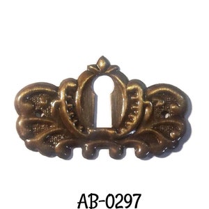 May include: A gold-toned keyhole cover with a decorative, ornate design. The cover has a keyhole in the center and a decorative flourish at the top. The text "AB-0297" is printed below the cover.