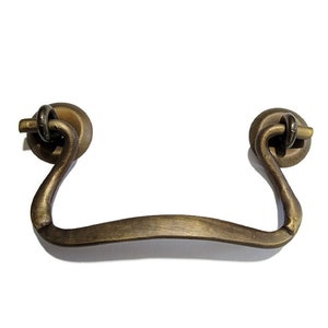May include: A brass drawer pull with a curved handle and rounded ends. The pull has a rustic, antique finish.