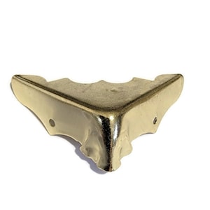 May include: Gold metal corner protector with a decorative design. The protector has a scalloped edge and a smooth finish.