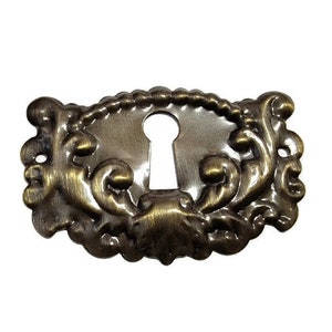 May include: A decorative keyhole cover made of brass with an ornate design. The cover has a keyhole in the center and is shaped like an oval.