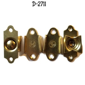 May include: Four brass-colored cabinet hardware pieces. The pieces include a ball catch, two plates with a circular emblem, and a spring-loaded catch. The text "D-2711" is printed in black at the top of the image.