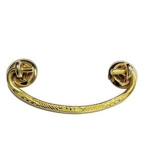 May include: A gold-toned metal drawer pull with a curved design and a decorative etched pattern. The pull has two round knobs on each end.