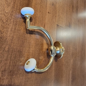 May include: A gold-toned coat hook with two white ceramic knobs. The hook is mounted on a wooden surface.