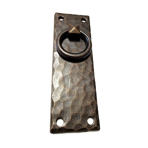 May include: A rustic, hammered metal door pull with a ring handle. The pull has a dark brown finish and is rectangular in shape.