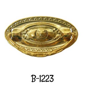 May include: A gold-toned oval-shaped cabinet pull with a decorative design and a ring handle. The pull has a raised center with a floral design. The text "B-1223" is printed below the pull.