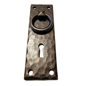 May include: A rustic, hammered iron door handle with a ring pull and a keyhole. The handle is a dark brown color and has a distressed finish.