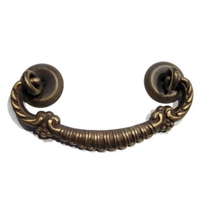 May include: A brass drawer pull with a decorative, ornate design. The pull has a curved shape and features a rope-like pattern and floral accents.