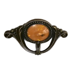 May include: Antique bronze-toned drawer pull with an oval amber-colored center. The pull features ornate scrollwork and a decorative design, suitable for furniture restoration or adding vintage charm.