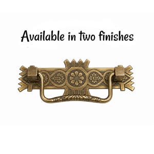 May include: A brass-toned metal drawer pull featuring an ornate rectangular backplate with a central flower motif and geometric edges. A U-shaped drop handle is attached to the plate. The text "Available in two finishes" appears above.