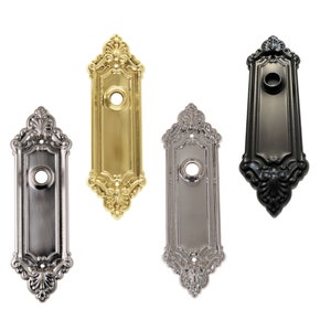 May include: Four ornate door plates in different finishes: gold, silver, nickel, and black. Each plate features a decorative design with a central hole for a doorbell or door knocker.