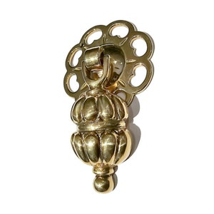 May include: A gold-toned cabinet pull with a decorative, ornate design. The pull features a round, fluted knob with a decorative finial and a decorative, openwork ring.