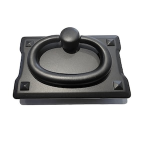May include: A black cabinet pull with a ring handle. The rectangular base has decorative corner accents. The ring handle is centered on the base and is attached to a small, round knob. The hardware is made of metal and has a matte finish.