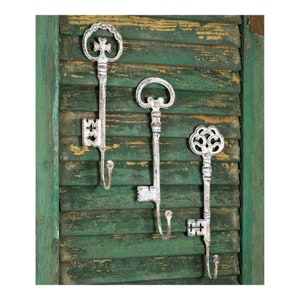 May include: Three decorative white metal key-shaped wall hooks. The hooks are mounted on a distressed green wooden shutter. Each key has a different ornate design, with a hook at the bottom for hanging items.