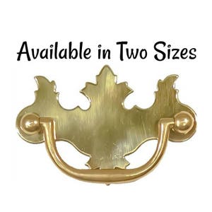 May include: A brass-colored drawer pull with a leaf-shaped backplate and a curved handle. The text "Available in Two Sizes" is above the pull. This hardware is suitable for cabinets and furniture.